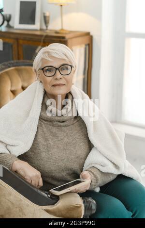 Die elegante alte Dame sitzt auf dem Lederarmstuhl und hält EIN Tablet in der Hand. Stockfoto