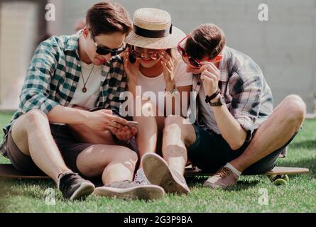 Drei junge Freunde Sitzen auf dem Rasen im Freien und Blick auf Handy. Stockfoto
