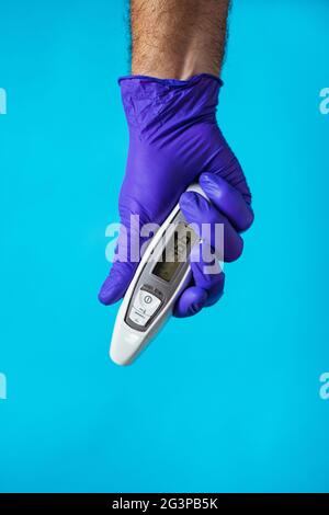 Die Hand eines Arztes trägt einen violetten Handschuh und hält ein Thermometer auf blauem Hintergrund. Corona-Virenschutz. Stockfoto