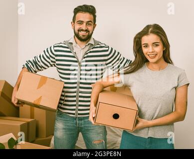 Junges Paar lächelt und hält Kisten, während es sich zwischen unverpackten Kisten aufhält Stockfoto