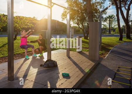 Personal Fitness Trainer und junge Frau Client, korrigiert Übungen mit Gurten. Morgens im Park. Sonniger Tag. Stockfoto