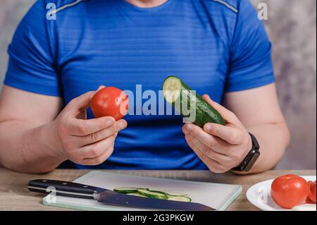 Ein Mann hält eine Gurke und eine Tomate in den Händen. Auf dem Brett befindet sich ein Messer und auf dem Tisch eine geschnittene Gurke Stockfoto