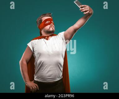 Super Hero Mann nimmt Selfie von Mobiltelefonen. Mann in EINEM Superhelden Kostüm in EINER roten Maske und Mantel isoliert auf Biskaya Green Rücken Stockfoto