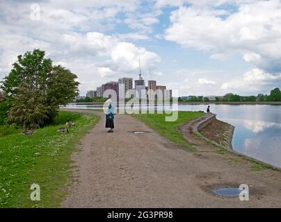 Ältere Frau, die auf dem Pfad steht und Gänse beobachtet, die sich am Stausee ernähren und Wohnwohnungen im Bau sind Walthamstow Wetlands UK DEWITT Stockfoto