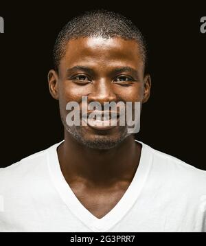 Lächelnder afroamerikanischer Mann, der die Kamera anschaut. Porträt eines dunkelhäutigen Mannes in weißem T-Shirt auf schwarzem Hintergrund. Nahaufnahme Stockfoto