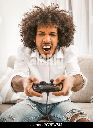 Schreiende Gamer schießt oder greift im Computerspiel an. Das Konzept der Emotionen. Junge Araber spielen Computerspiel sitzen auf dem Sofa in Stockfoto