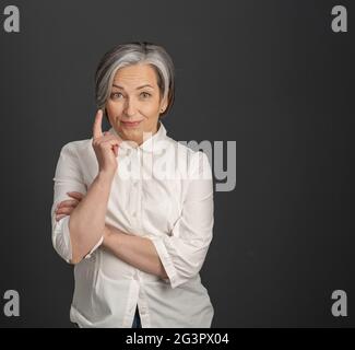 Haben Idee Geste von reifen Frau. Die grauhaarige Geschäftsfrau hob ihren Zeigefinger nach oben, während sie die Kamera auf grau posierte Stockfoto