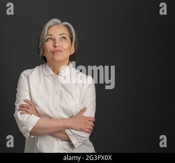 Kreative reife Frau, die mit gekreuzten Armen nachdenklich nach oben blickt. Charmante silberhaarige Frau lächelt zärtlich auf dunklem g Stockfoto