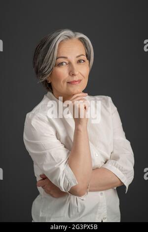 Die wunderschöne grauhaarige Frau lächelt sanft, während sie ihr Kinn mit der Hand berührt und auf die Kamera schaut. Intelligent schön reif Stockfoto