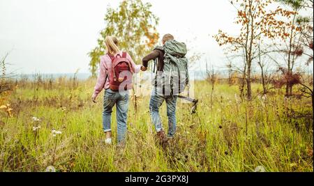 Rückansicht eines Reisenden Paares von Rucksacktouristen, die im Freien durch ein Herbstfeld watten und Hände mit Wanderstöcken hielten. Wandern Co Stockfoto