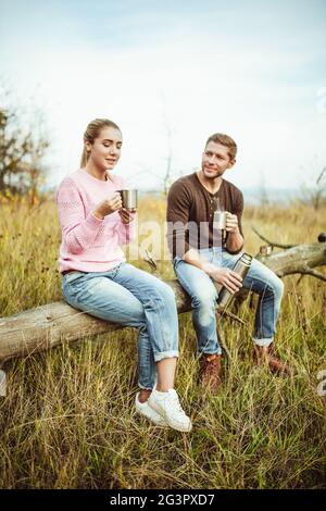 Tee-Party oder Kaffee trinken im Freien. Ein fröhliches Paar trinkt heißen Kaffee oder Tee und kommuniziert auf einem Holzbalken im Freien Stockfoto