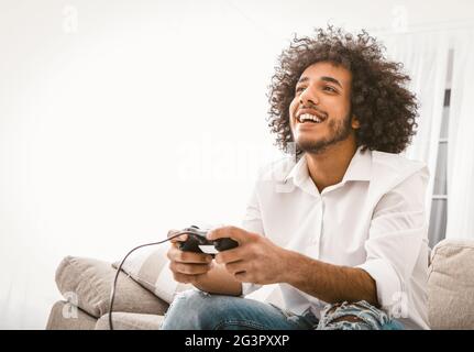 Glücklicher Mann, der Computerspiele spielt. Der junge arabische Kerl unterhält sich zu Hause in Selbstisolation während der Quarantäne. Platz auf der linken Seite kopieren Stockfoto