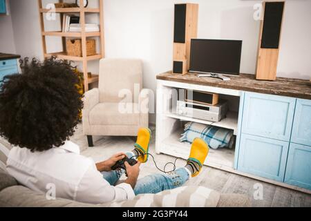 Stilvoller Kerl spielt Computerspiele, die auf der Couch sitzen. Junger unkenntlich Mann mit afro Frisur in gelben Pantoffeln entspannen in h Stockfoto