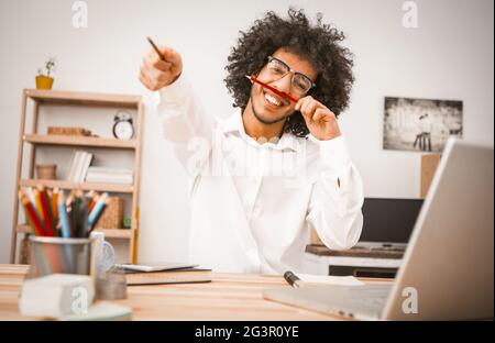 Fröhlicher Schüler, der während der Arbeitspause Spaß hat. Der junge Araber im weißen Hemd lächelt breit und streicht mit Bleistiften in den Händen. Stockfoto