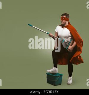 Superheld hat Spaß mit Reinigungsgeräten. Mann in rotem Umhang hält Mopp und präsentiert sich als Musiker mit Gitarre. Isoliert auf g Stockfoto