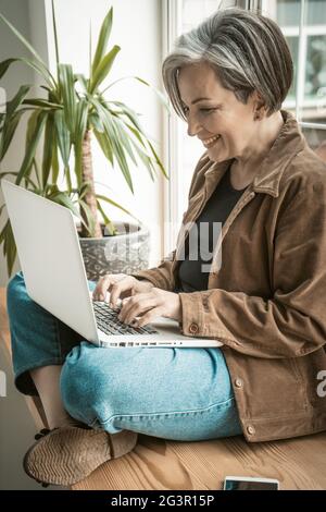 Glückliche, reife Frau, die auf dem Laptop chattet und sich gegen das Fenster lehnt. Pretty Lady lächelt am Computer. Seitenansicht. Zinn Stockfoto