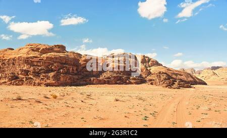 Felsige Massive auf roter Sandwüste, Fahrzeugspuren Boden, heller wolkiger Himmel im Hintergrund, typische Landschaft im Wadi Rum, Jordanien Stockfoto