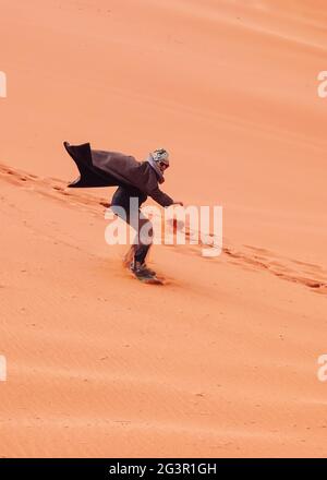 Junger Mann, der Sanddüne surft, trägt bisht - einen traditionellen Beduinenmantel. Sandsurfing ist eine der Attraktionen in der Wadi Rum Wüste Stockfoto