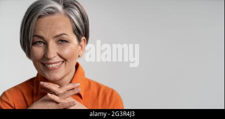 Reife Frau mit gekreuzten Daumen breit lächelnd. Fröhliches weibliches Model in orangefarbenem Hemd. Nahaufnahme im Hochformat. Reifegradkonzept. Hori Stockfoto