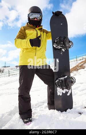 Snowboarder Mann mit Snowboard steht auf Berghang, hält sein Brett, trägt eine verspiegelte Brille, Daumen nach oben Stockfoto