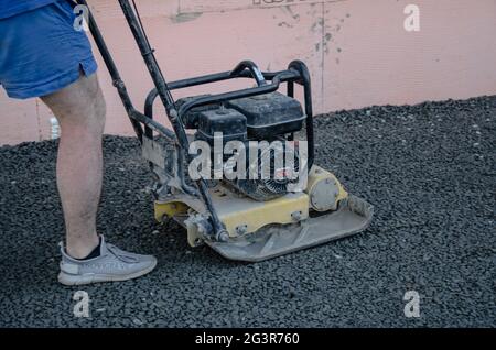 Ein Straßendienstmitarbeiter verdichtet den Asphalt auf einem eingezäunten Straßenabschnitt der Fahrbahn mit einem Vibrationsplatten-Verdichter. Stockfoto