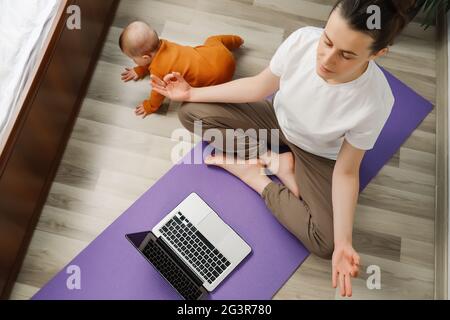Junge Mutter tragen Activewear Sitzen Sie auf der Matte in Lotusposition dreht sich auf Laptop entspannende Musik für Stressabbau Meditationspraxis in der Nähe verspielten niedlichen BA Stockfoto
