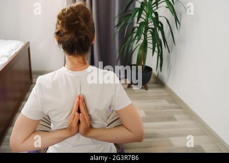 Junge Frau praktiziert Yoga, macht Namaste hinter dem Rücken, arbeitet, allein zu Hause. Fit gesunde aktive Mädchen sitzen auf Matte auf dem Boden im Wohnzimmer, r Stockfoto