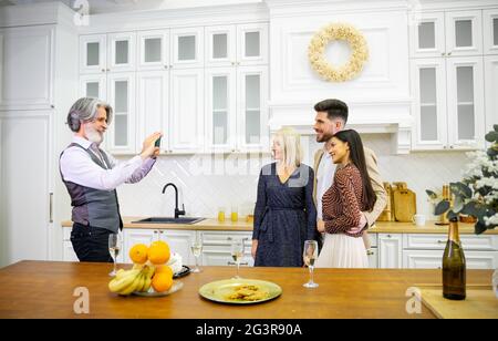 Eleganter älterer grauhaariger Mann Großvater, der während der Geburtstagsfeier seine Frau und sein junges Paar fotografiert, die in einer stilvollen Küche zu Hause mit Obst, Süßigkeiten und Champagner auf dem Tisch stattfindet Stockfoto