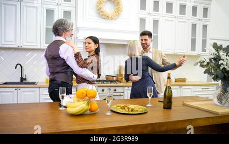 Glückliche Familienmitglieder tanzen während der Geburtstagsfeier in der Küche neben dem Tisch mit Kuchen und Champagner zu Hause, Großeltern und junge verheiratete Paar Spaß haben und genießen Hausfeier Stockfoto