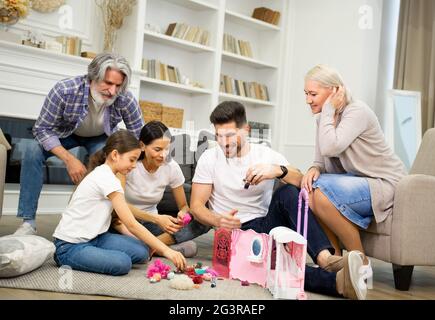 Große glückliche, generationenübergreifende Familie Vater Mutter und ältere Großeltern, die aufgeregt sind, während sie mit einem entzückenden kleinen Mädchen spielen, das ein neues Puppenhaus zeigt, und eine tolle Zeit zusammen zu Hause genießen Stockfoto