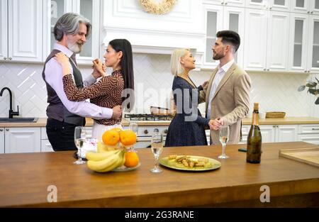 Familienfeier. Die junge Tochter tanzt mit dem älteren Vater, während ihr gutaussehender Mann während der Geburtstagsfeier mit seiner Schwiegermutter in der Küche zu Hause mit Kuchen und Champagner tanzt Stockfoto