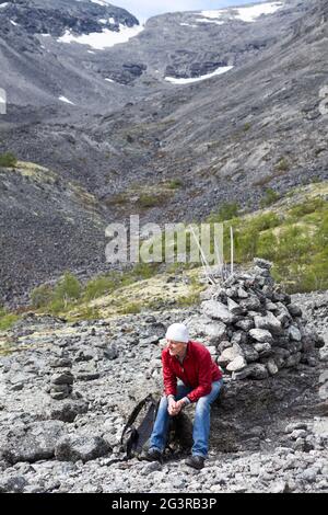 Einsamer kaukasischer reifer Rucksacktourist, der auf Stein sitzt und auf dem Hintergrund zum Bergpass klettert Stockfoto