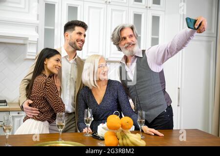 Positive Großeltern und junge Paare in formeller Kleidung, die Selfie machen oder Videoanruf auf dem Smartphone machen, die Kamera betrachten und während der Feiertagsfeier mit der Familie zu Hause lächeln Stockfoto