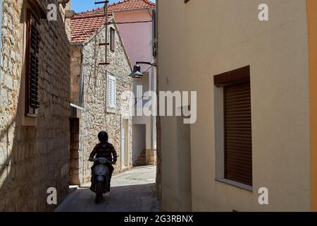 Ein Fahrer auf einem Roller, der eine enge Steinstraße im Dorf Jelsa auf der Insel Hvar, Kroatien, hinunterfährt Stockfoto