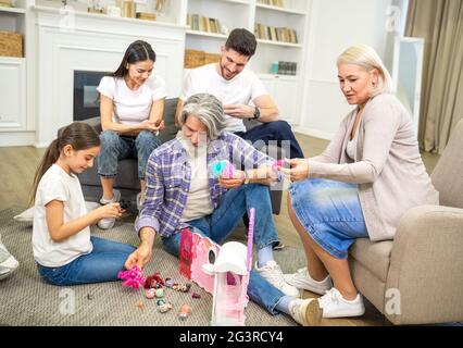 Große glückliche Mehrgenerationenfamilie Vater Mutter und Großeltern spielen mit niedlichen kleinen Mädchen Enkelin, Blick auf neue Puppenhaus, Spaß haben und zusammen am Wochenende zu Hause entspannen Stockfoto