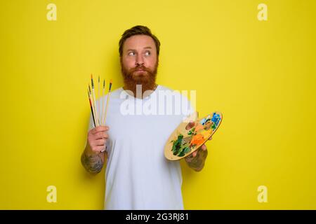 Unentschloßter Mann mit Bart und Tattoo ist bereit, mit Pinsel zu zeichnen Stockfoto