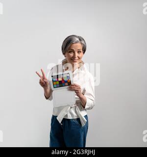 Frau mit grauen Haaren hält colorchecker auf einem isolierten Hintergrund. Erwachsene Frau lächelt und zeigt zwei Finger. Hochwertige Fotos Stockfoto