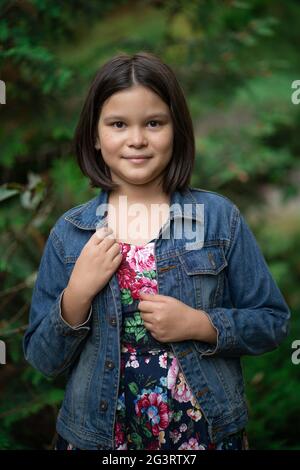 Nettes kleines Mädchen trägt karierten Kleid und Denim Jacke posiert Vor dem Hintergrund der grünen Natur Stockfoto