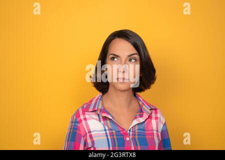 Denkende junge Frau trifft ernsthafte Entscheidung. Hübsche Brünette schaut nach oben auf den Kopierplatz auf der linken Seite. Isoliert auf gelbem Hintergrund Stockfoto