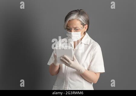 Ärztin arbeitet mit digitalen Tabletten. Kaukasische Frau in Schutzmaske und Handschuhen steht gegen graue Wand auf dem Hintergrund Stockfoto