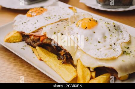 Rindersteak mit Schinken, Käse und zwei Spiegeleiern, begleitet von französischen Speisen Stockfoto