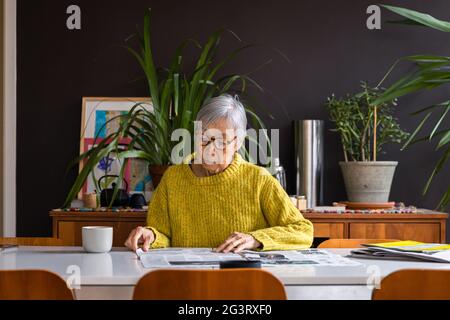 Ältere Frau mit gelbem Pullover, die Zeitung liest, während sie am Esstisch sitzt Stockfoto
