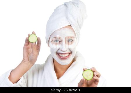 Junge Frau im Bademantel, mit Gesichtsmaske, weißem Handtuch auf dem Kopf und Gurkenscheiben in den Händen Stockfoto