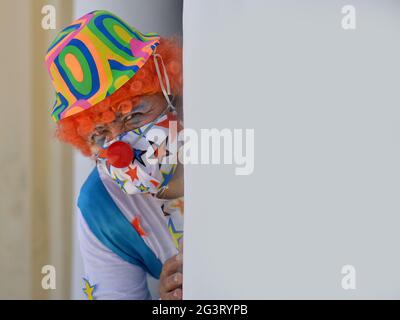 Männlicher Clown mit oranger Perücke, im Clown-Outfit und farbenfroher Stoffmaske gucken während der globalen Coronavirus-Pandemie um eine Ecke. Stockfoto