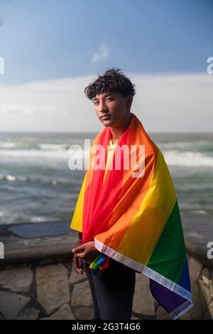 Der junge schwule Mann hält stolz die lgbt-Flagge in den Händen vor dem Meer, inklusive Stockfoto