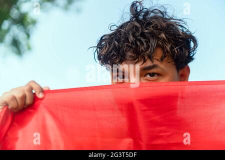 Ein junger schwuler Mann bedeckt einen Teil seines Gesichts mit der lgbt-Flagge und schaut in die Kamera Stockfoto