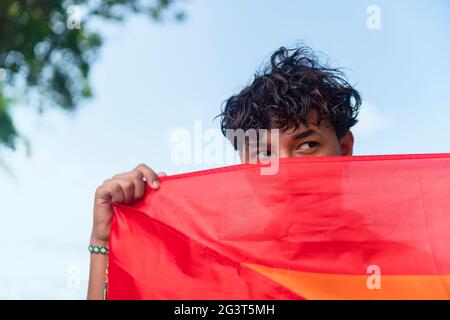 Ein junger schwuler Mann bedeckt einen Teil seines Gesichts mit der lgbt-Flagge und schaut in die Kamera Stockfoto