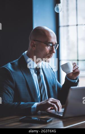 Ein respektabler Geschäftsmann trinkt Kaffee und arbeitet mit einem Computer vor dem Fenster. Seitenansicht des gut aussehenden kaukasischen schönen Typi Stockfoto