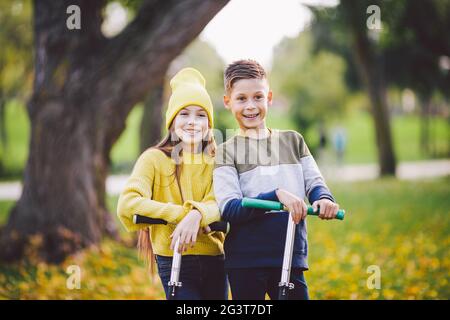 Zwillinge 10 Jahre alt in hellen Kleidern verbringen Zeit aktiv zusammen im Herbstpark Reiten Kick Scooter. Frohe Kinder. Eco-Übertragung Stockfoto
