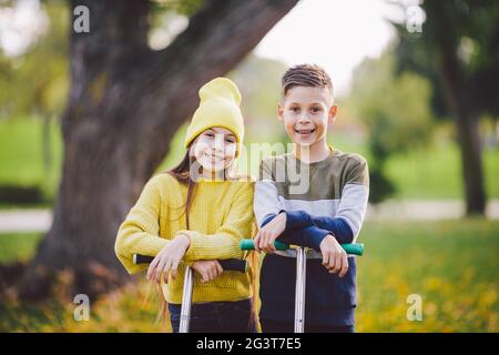 Zwei Kinder kaukasischen Jungen und Mädchen Zwillinge posieren mit Motorroller im Herbst Park. Glückliche Kinder reiten Kick Scooter im öffentlichen Park. K Stockfoto